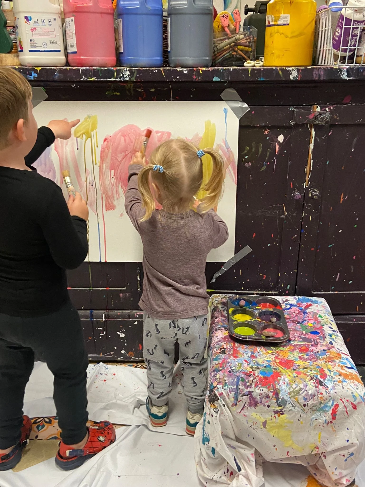 Two children painting on a large white canvas, with paint bottles on a shelf above and a paint palette on a splattered cloth-covered table nearby.