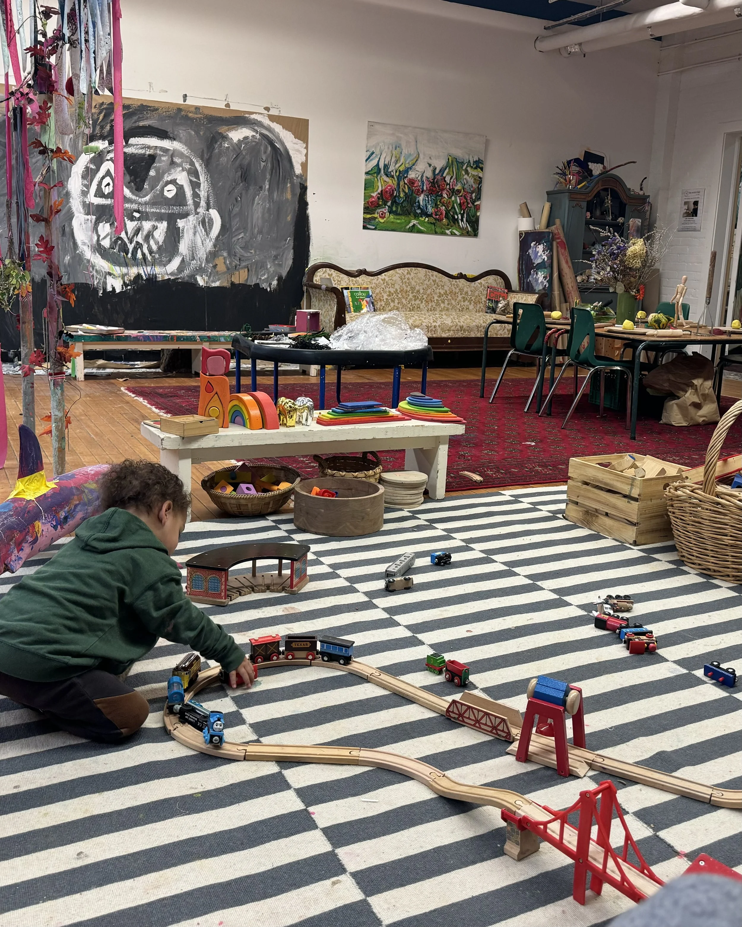 A young child in a green hoodie playing with a wooden toy train set on a black and white striped carpet in a colorful and artistic room filled with furniture, artwork, and craft supplies.