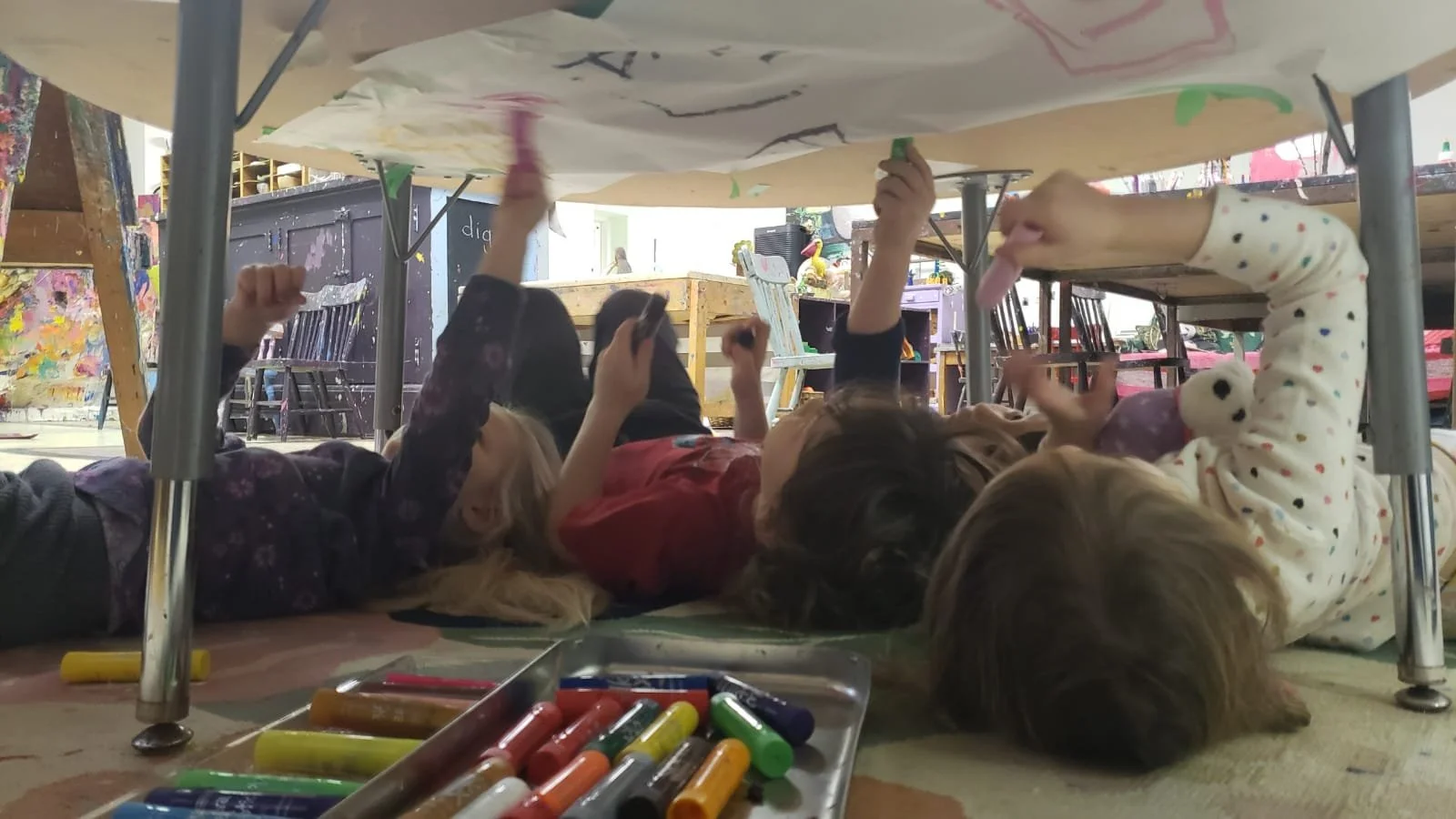 Four children lying on the floor under a table, drawing and coloring with markers and crayons.