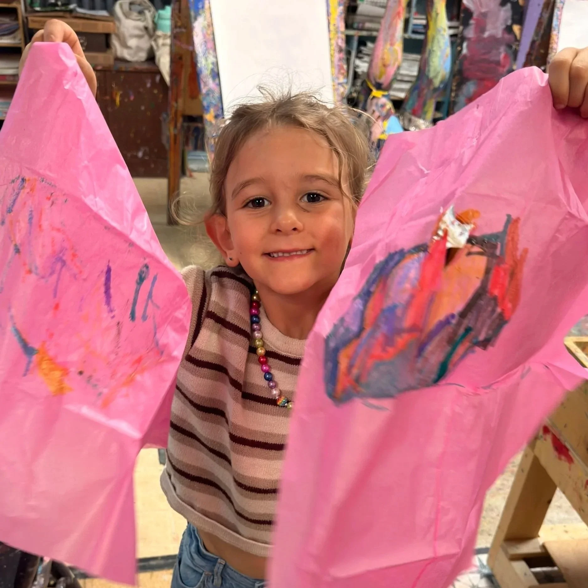 Preschool child holding up two pink tissue paper process art pieces with oil pastel marks in The Artful Child studio