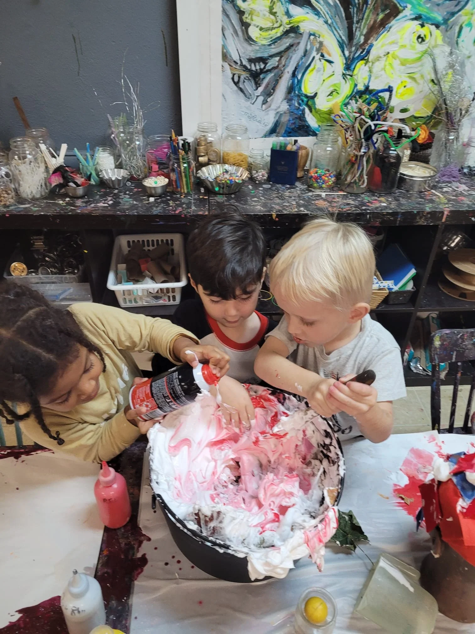 Three children are making a craft project with foam and marshmallow in a craft space with jars and art supplies on a black, paint-splattered table, with colorful abstract artwork hanging in the background.