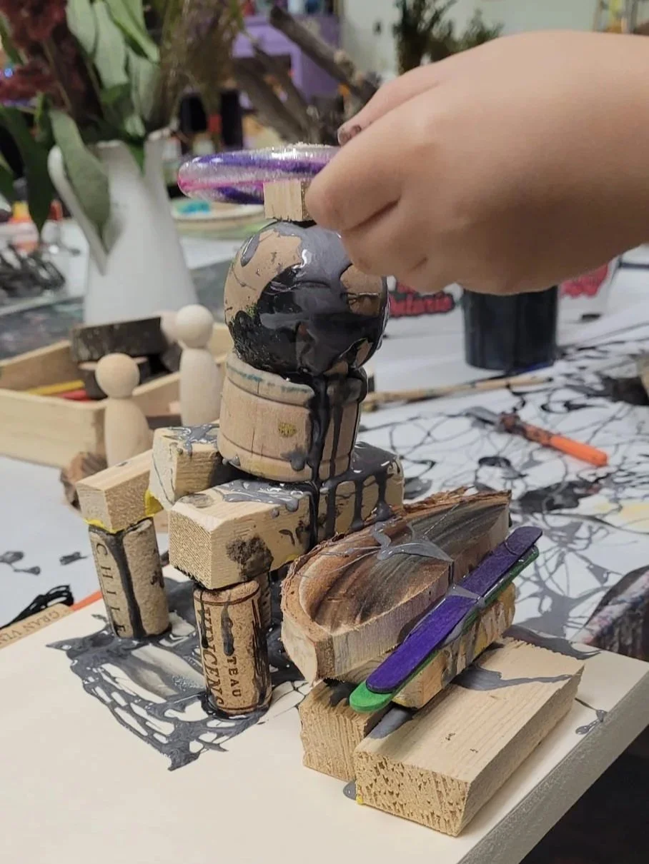 A hand pouring black paint onto a small structure made of wooden blocks, wine corks, and a piece of wood, with some paint drips visible.