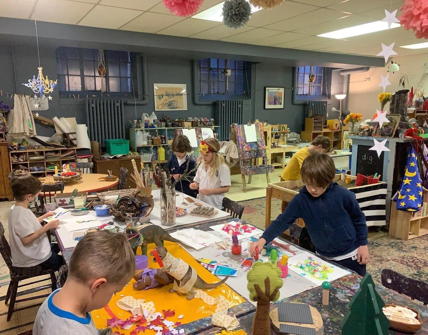 Children participating in arts and crafts activities in a creative classroom or art studio, with various art supplies, colorful decorations, and paintings visible.