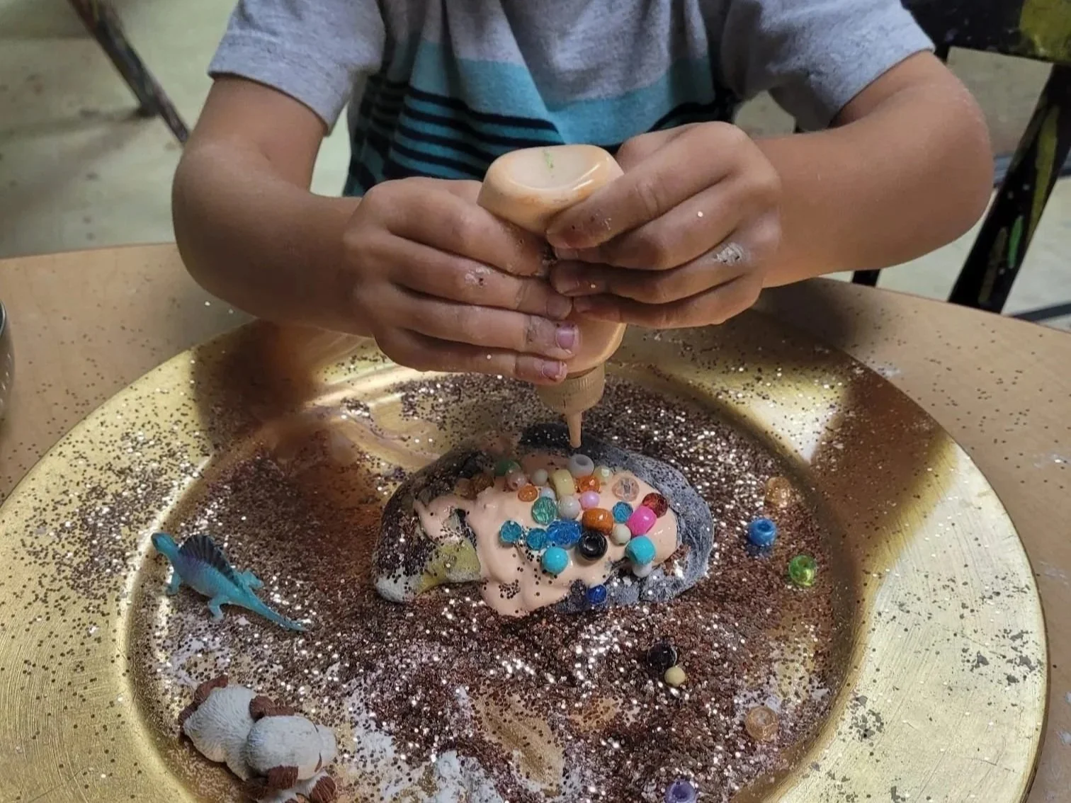 Child sculpting and decorating a small model with colorful beads and glitter on a golden tray.