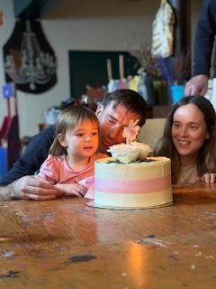 A man is blowing out birthday candles on a cake with two women and a young girl watching, in a cozy indoor setting.