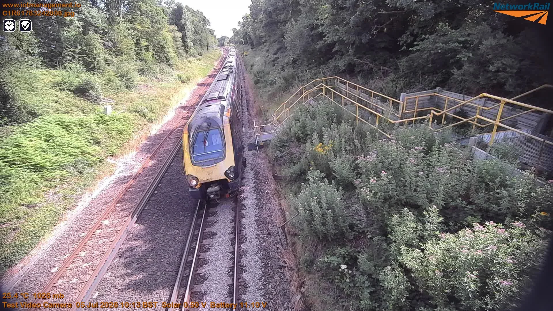 A yellow and black train traveling on railway tracks surrounded by lush green trees and bushes, with a staircase and railing to the right side of the image.