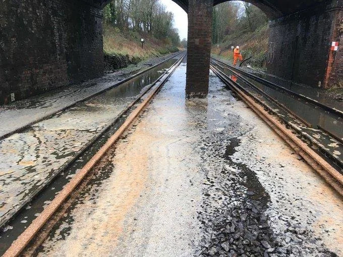 Flooded railway tracks under a bridge with a person in an orange safety vest in the distance.