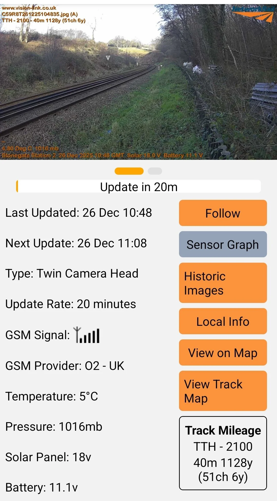 A photograph of rural train tracks with surrounding grass and trees, some fencing, and a traffic sign in the distance. Overlaid text includes weather and technical details, along with options to view sensor data, historic images, and maps.