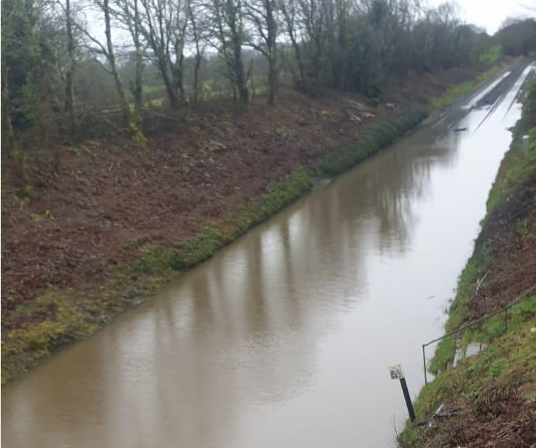 A canal or waterway with muddy water, lined by grassy slopes and leafless trees, with a freeway on the opposite bank during overcast weather.