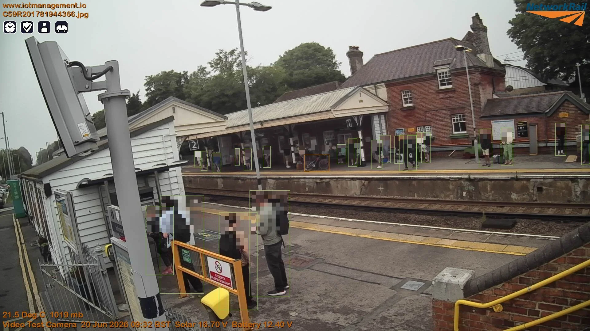 A train station platform with several people waiting and standing, a small white wooden shed, a train timetable board, and a historic brick building with chimneys in the background. Overhead sky and street lamps are also visible.