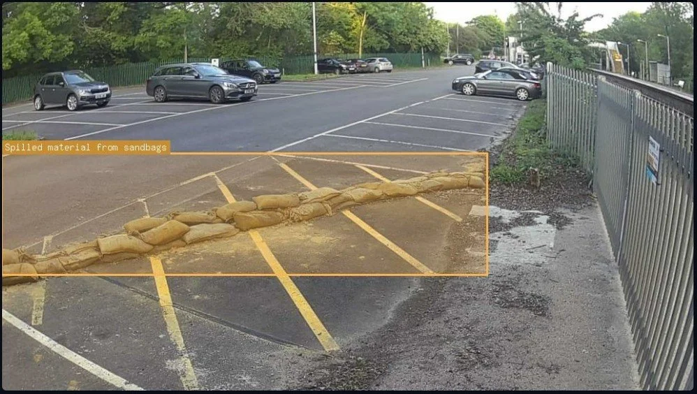 A parking lot with several parked cars and a pile of sandbags blocking part of the entrance, with an overlay indicating spilled material from sandbags.