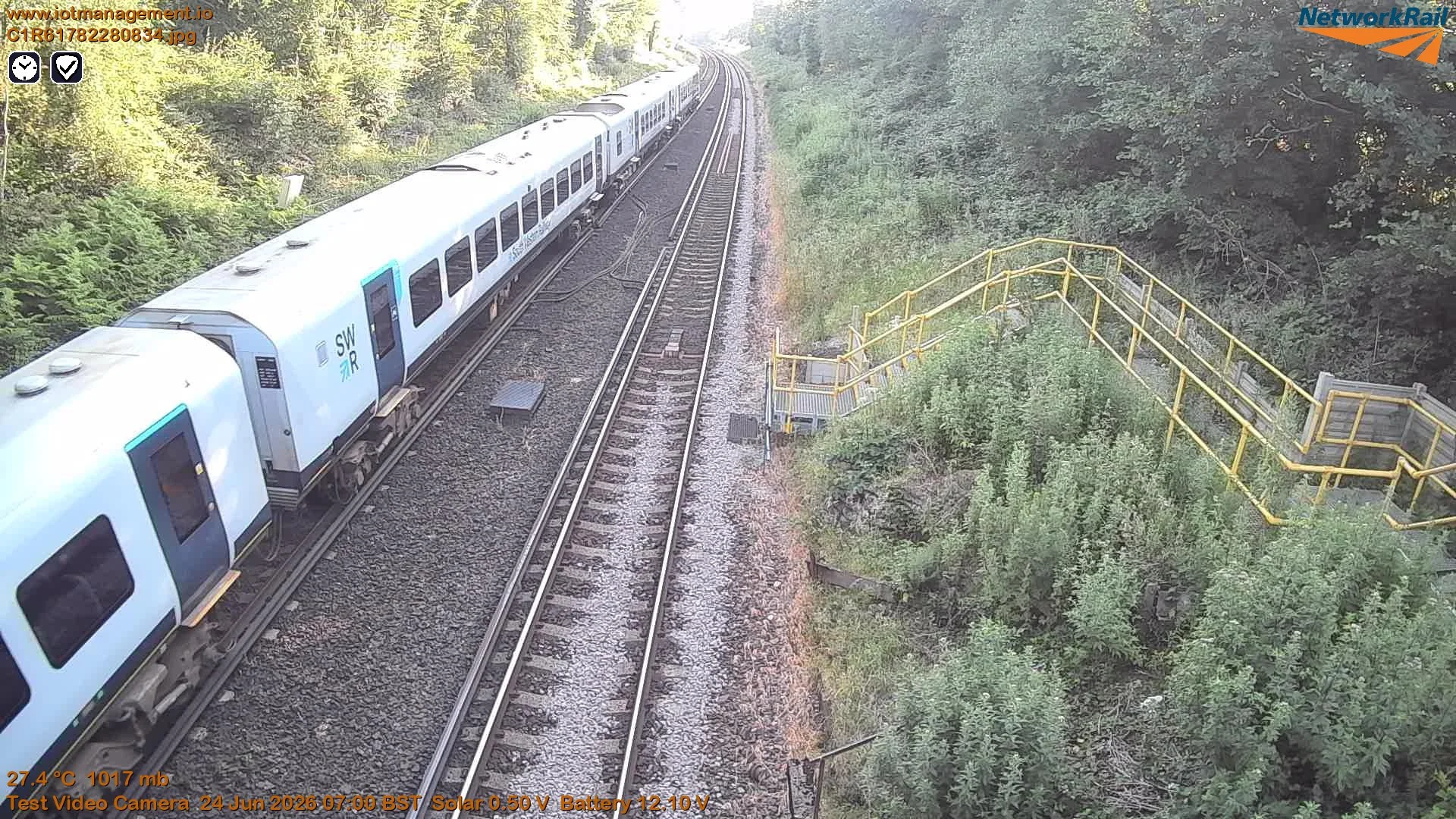A white passenger train on railway tracks surrounded by greenery and trees, with a yellow safety railing on the right side.