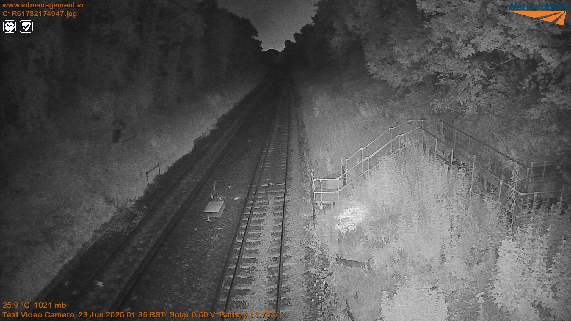 Nighttime view of a railway track running through a wooded area, captured by a test video camera, with some infrastructure and trees on either side.