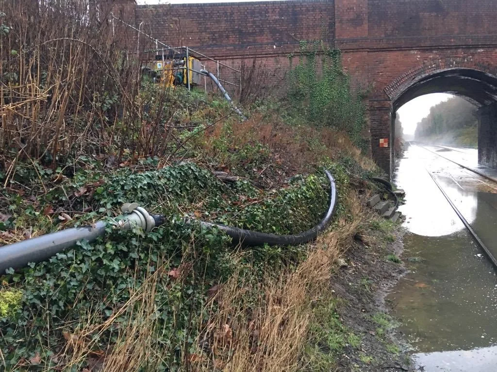 A pipeline leaks oil runoff into a creek next to railroad tracks passing under a brick tunnel arch.