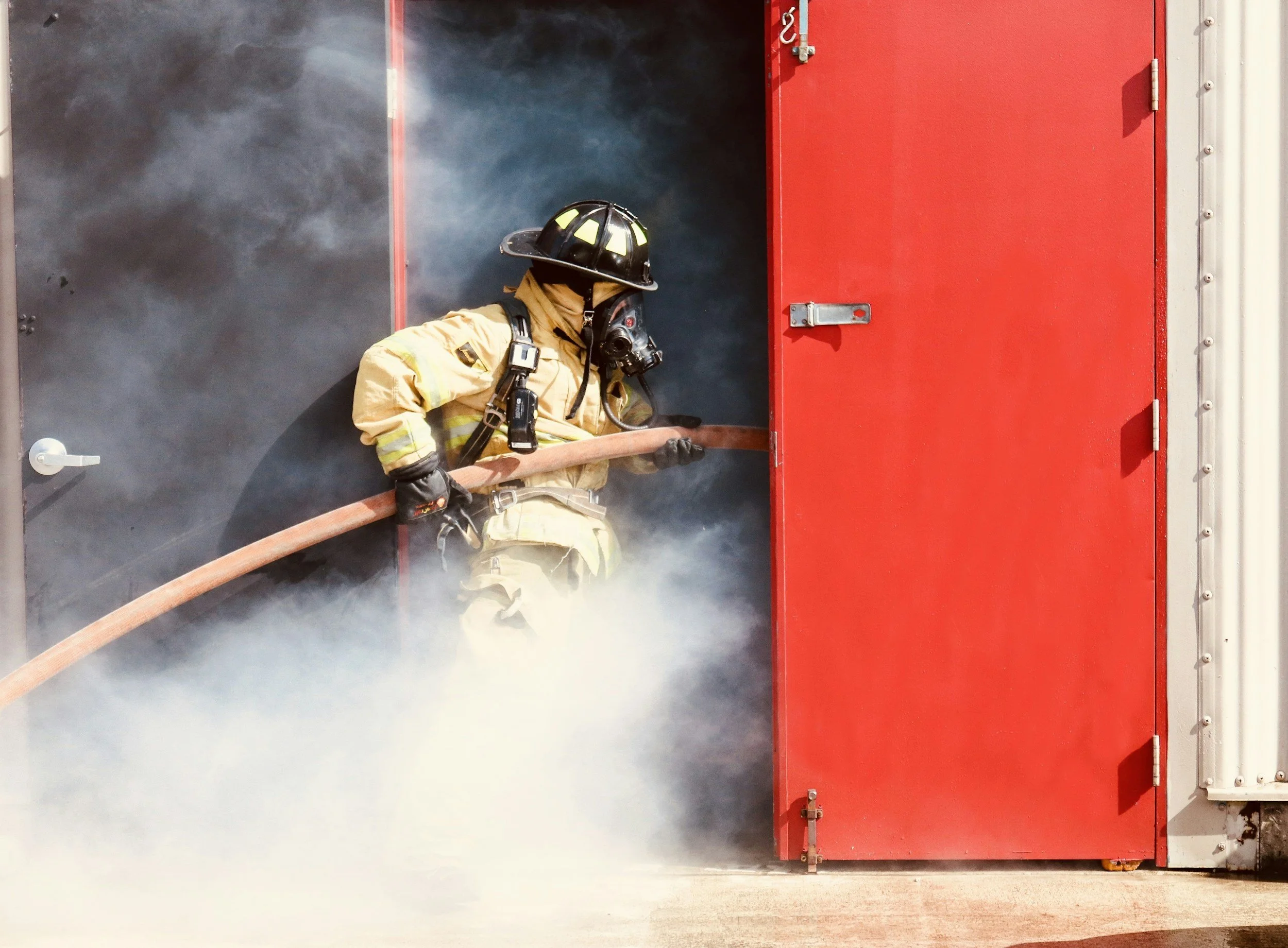 Firefighter wearing protective gear and helmet, using a hose to extinguish a fire inside a building with a red door.