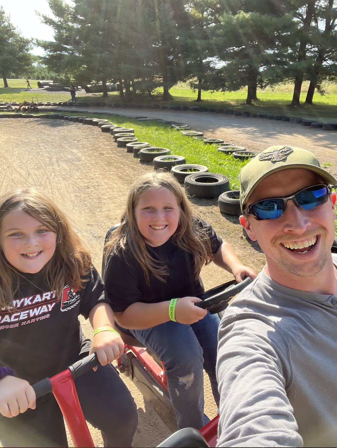 A man and two girls smiling and posing for a selfie at a go-kart track with tires lining the track and trees in the background.