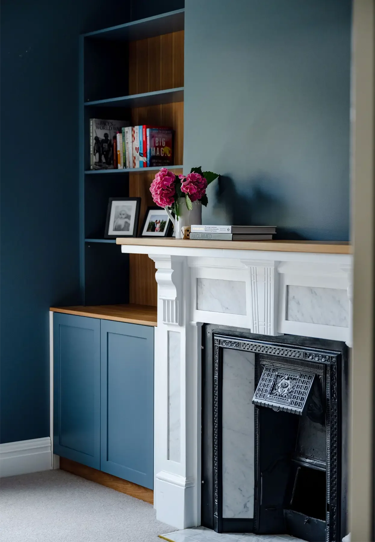 A living room corner with a white fireplace featuring a marble surround, a black fireplace insert, and a white mantel. A vase of pink hydrangeas and some books sit on top of the mantel. To the left, a blue cabinet with closed doors, and above it, a built-in bookshelf with various books and framed photos.