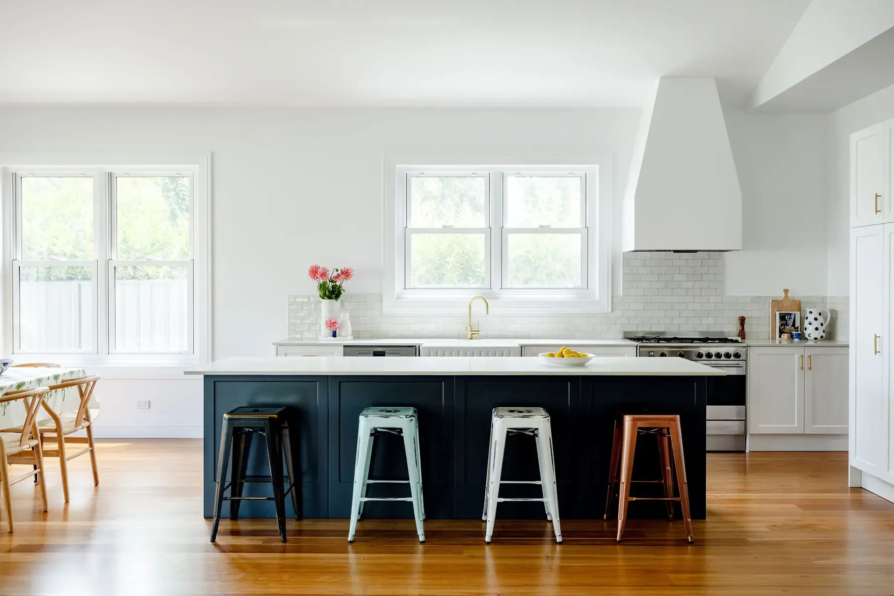 Kitchen island bench with a white benchtop and deep, deep blue cabinetry underneath. There are four rustic, metal stools in front, with a which kitchen bench in the background featured gold tapwear