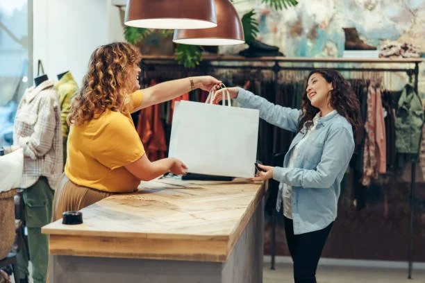 A woman with curly hair and a yellow shirt hands a shopping bag to another woman with long dark hair and a blue jacket in a clothing store.