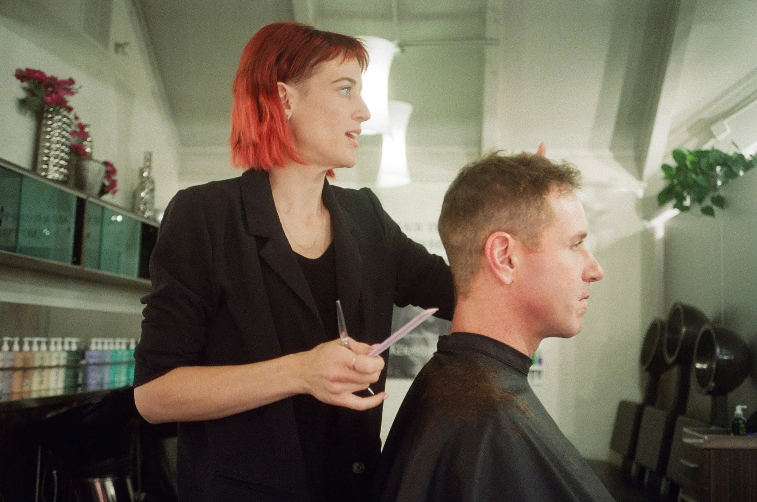 A hairstylist with red hair and black attire cutting a man's hair in a salon suite.