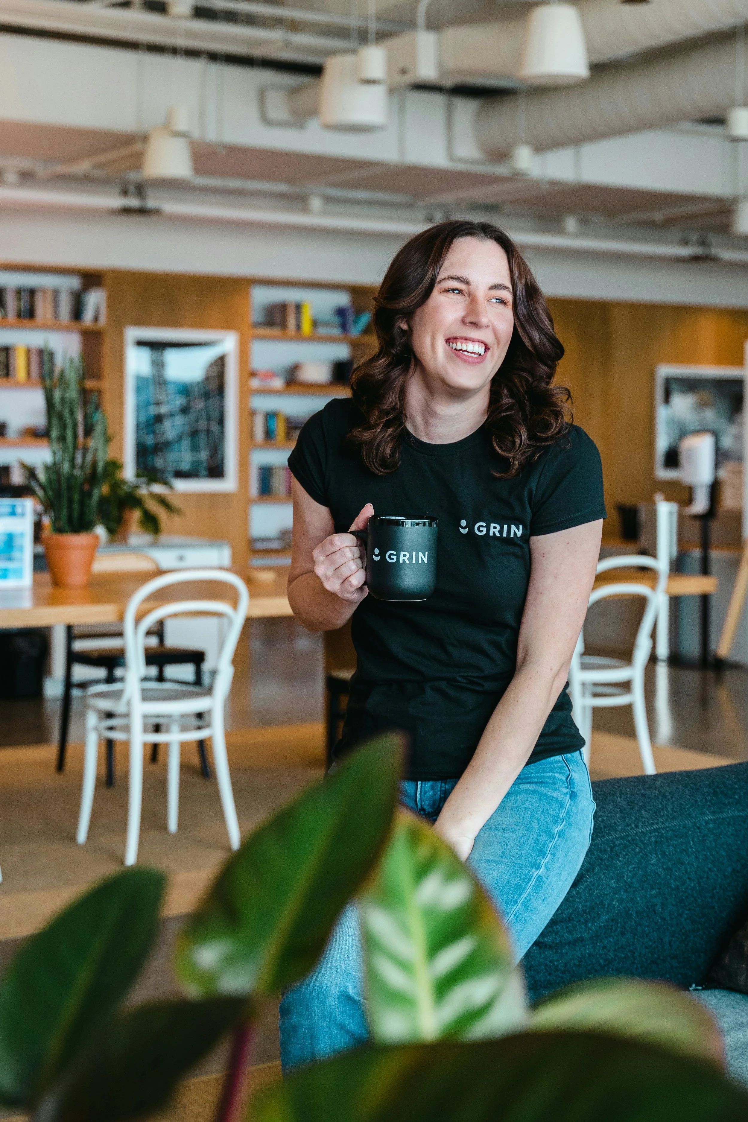 A woman with brown hair smiling and holding a black mug with the word "GRIN" on it in a modern office space.