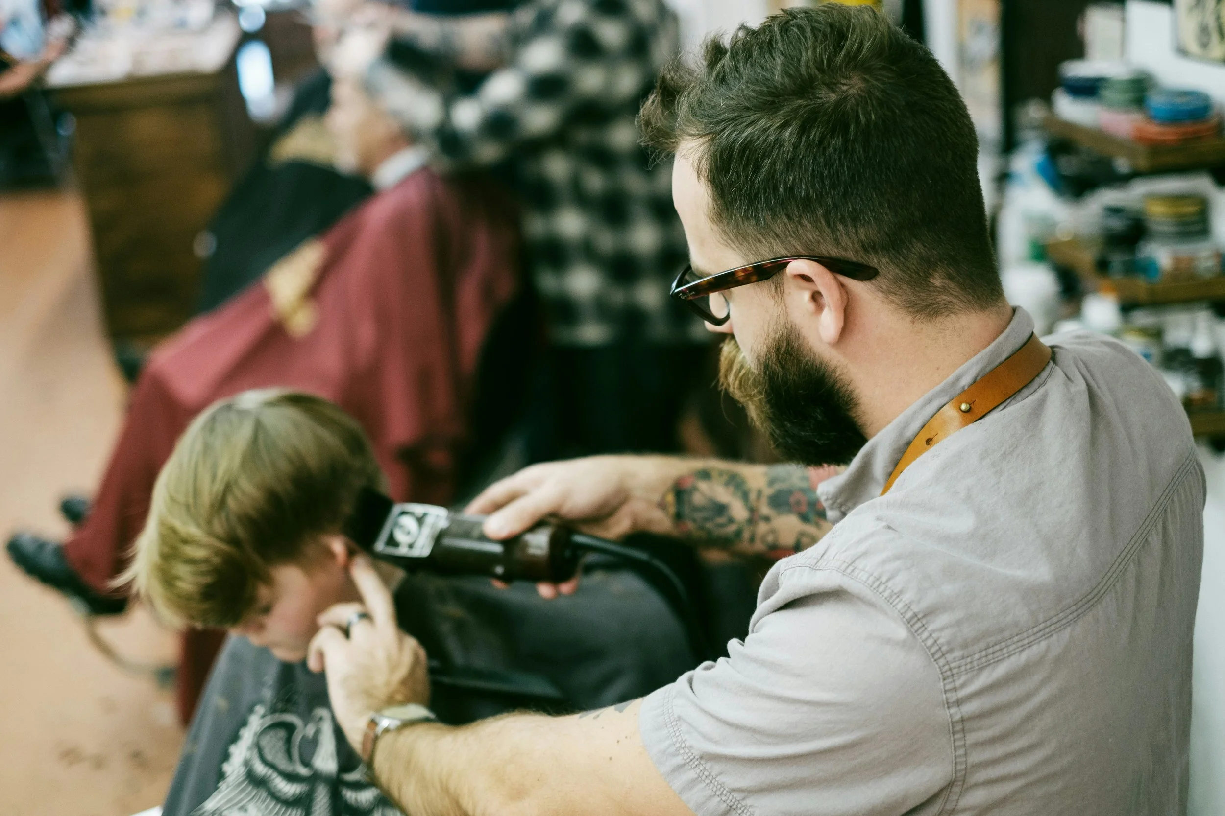 A man who leases a studio space inside of a salon suite business with tattoos, glasses, and a beard is in a barbershop using clippers to haircut a young boy with light brown hair, while other customers are in the background.