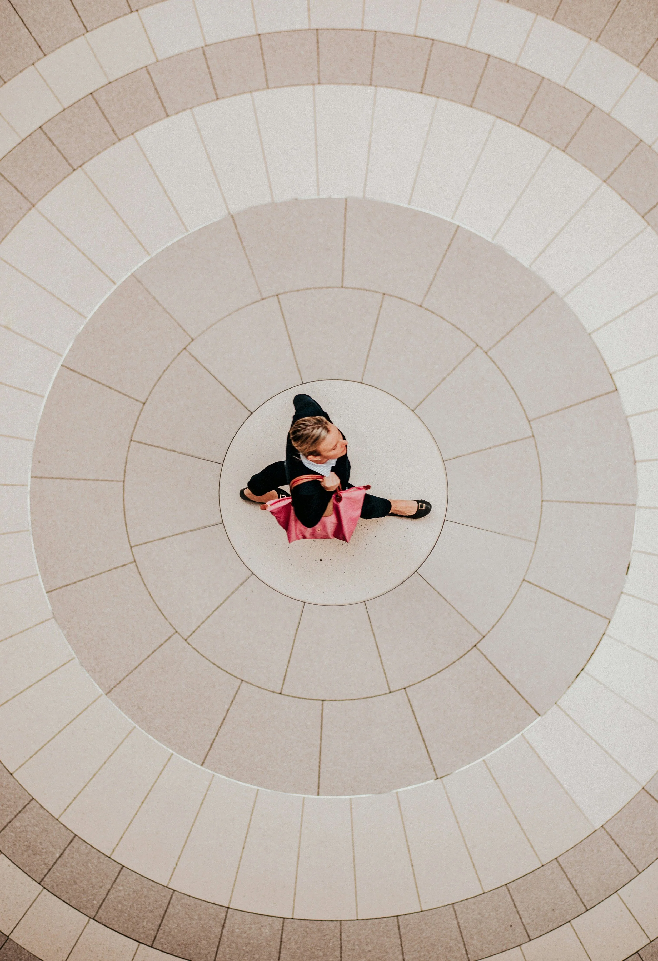 A woman with light brown hair, wearing a black jacket and black shoes, walking on a spiral staircase with beige and brown tiles, carrying a pink bag.