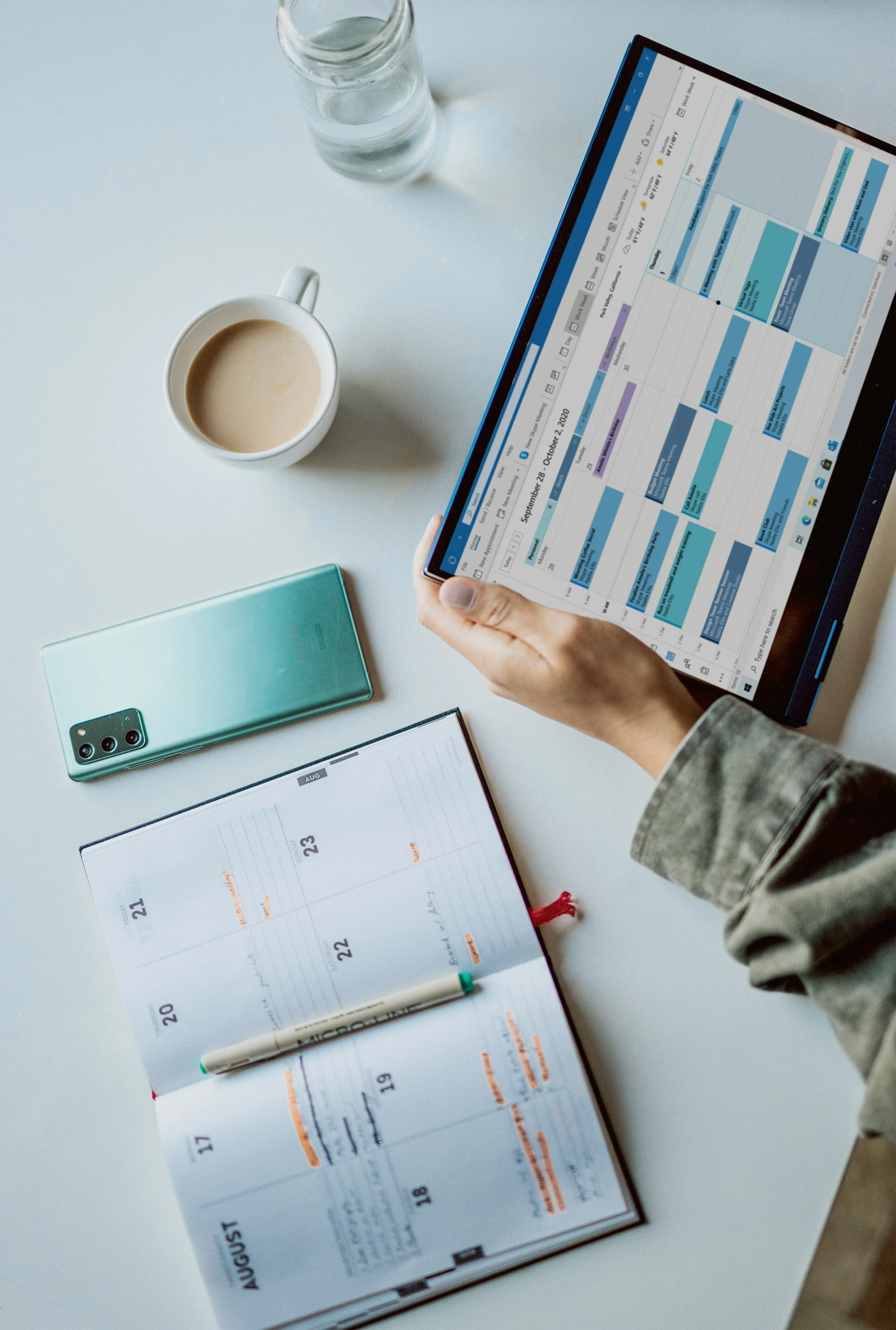 Person holding a digital tablet with a calendar displayed on it, surrounded by a coffee mug, a smartphone, an open planner, and a glass of water on a white table.