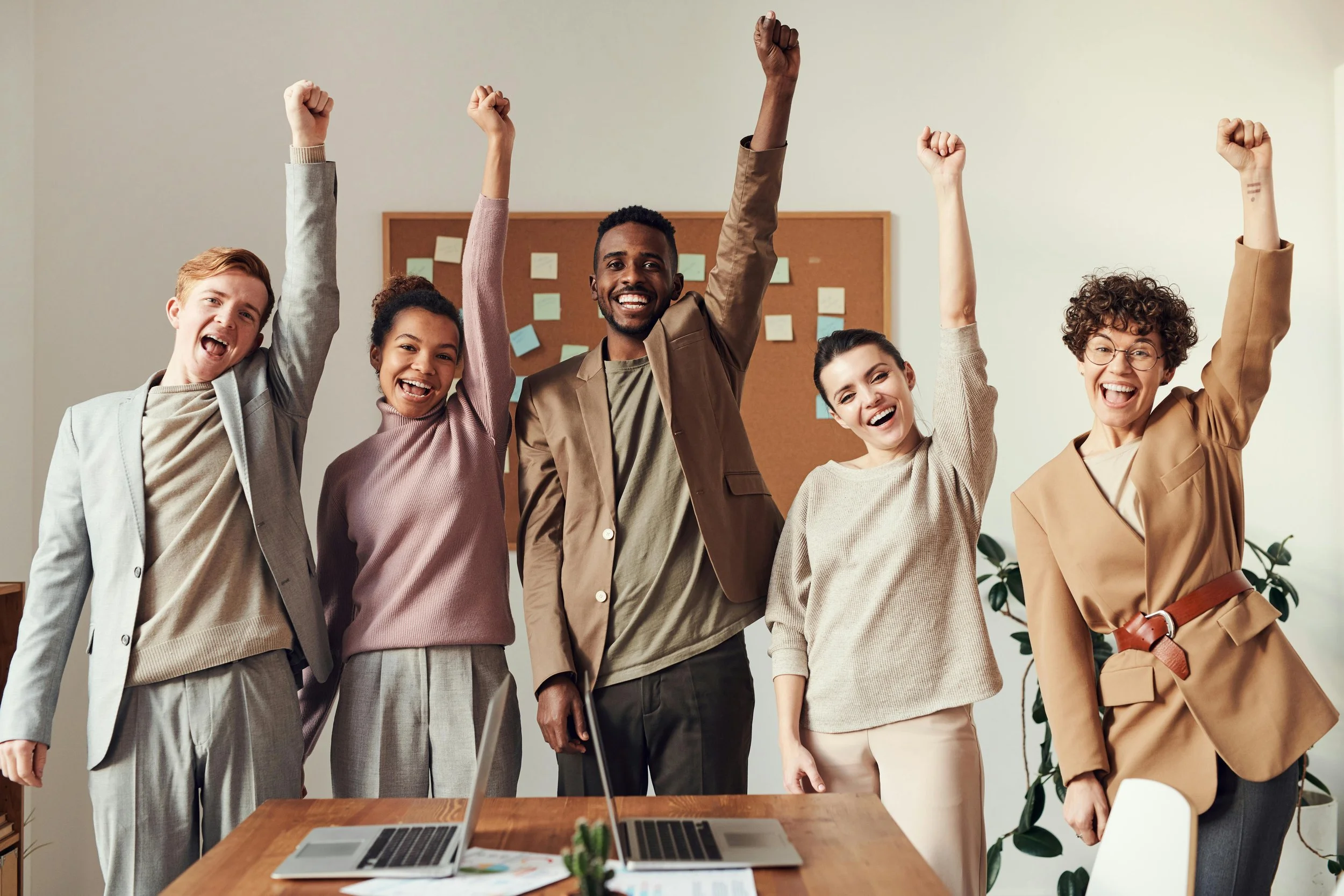 Group of five diverse young adults standing together in an office, smiling and raising their fists in celebration.
