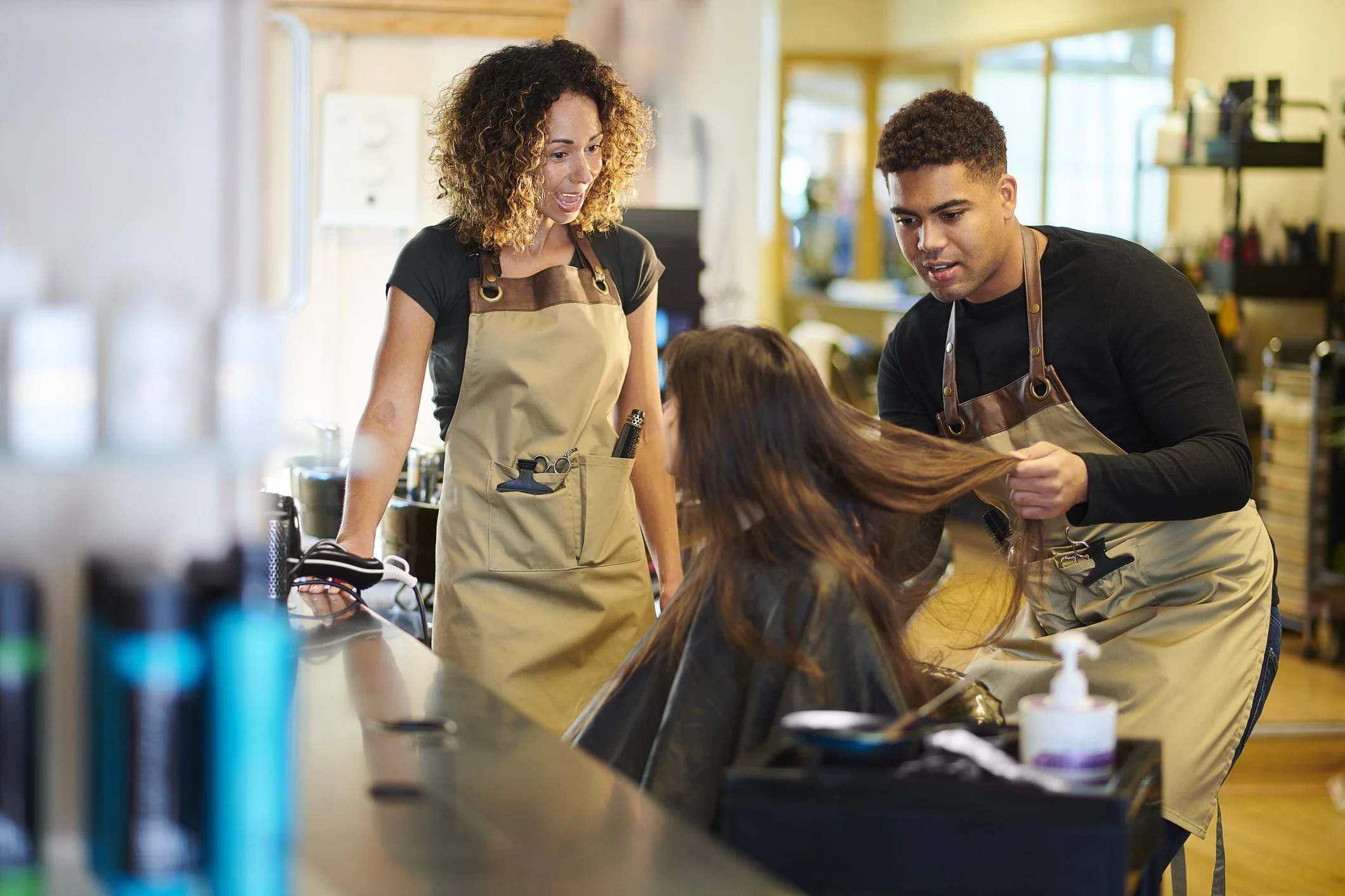 A woman with curly hair sitting at a salon chair. Two hairstylists are working on her hair, one holding a section of her long brown hair, both wearing beige aprons in a well-lit salon.