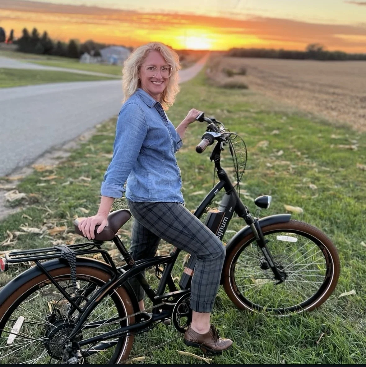 A woman with curly blonde hair and glasses, wearing a denim shirt and plaid pants, smiling while sitting on a black electric bicycle on a grassy area during sunset.