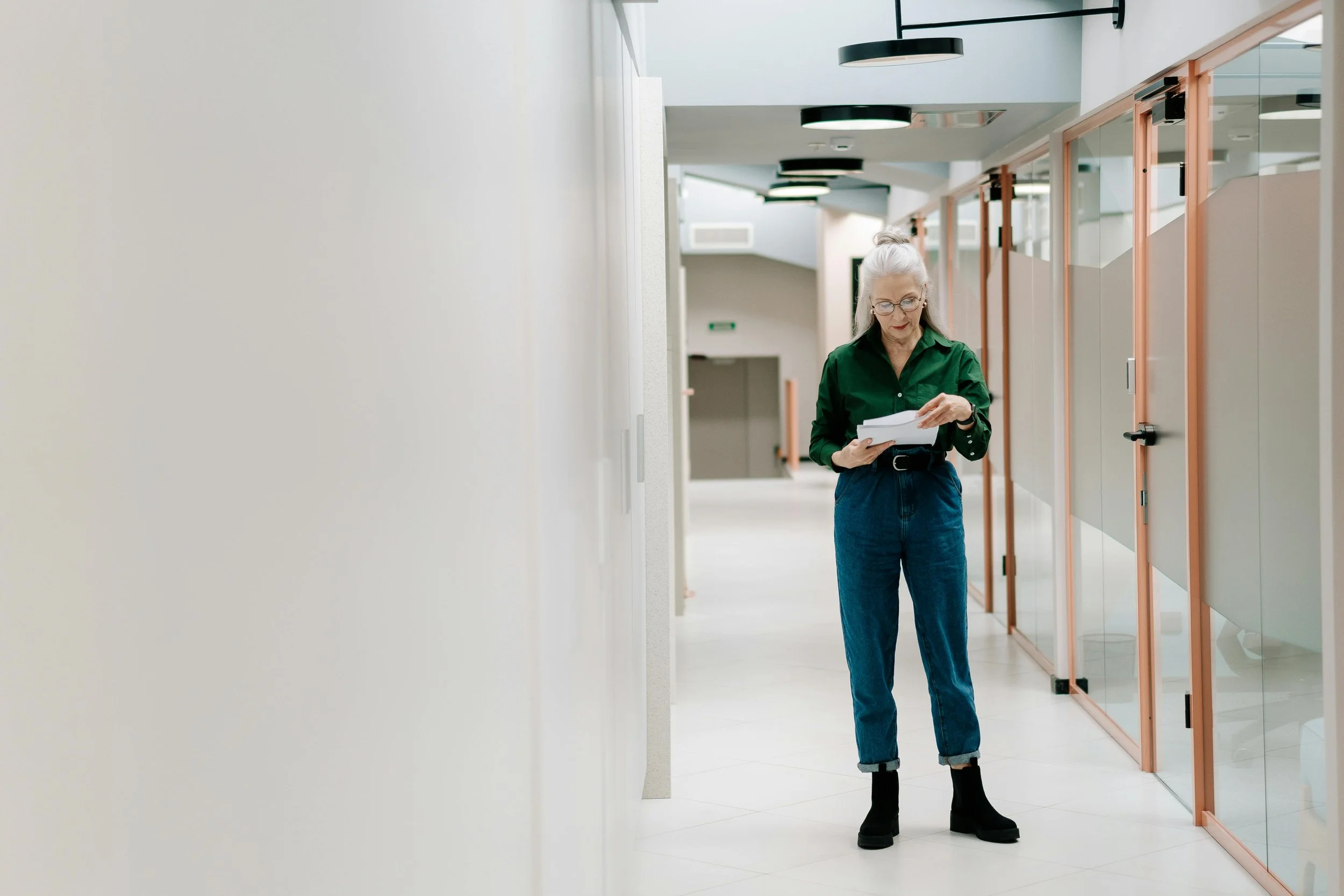 An elderly woman who owns a salon suite concept with silver hair tied in a bun, wearing glasses, a green shirt, blue jeans, and black boots, is standing in a modern, well-lit corridor reading a document about leasing space to beauty professionals.