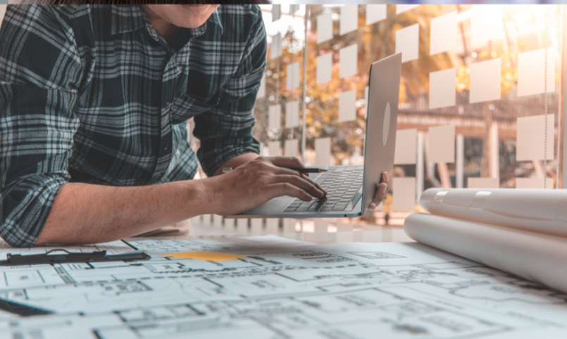 A person in a plaid shirt working on a laptop at a desk with architectural plans, a clipboard, and rolls of blueprints, with sunlight coming through a window.