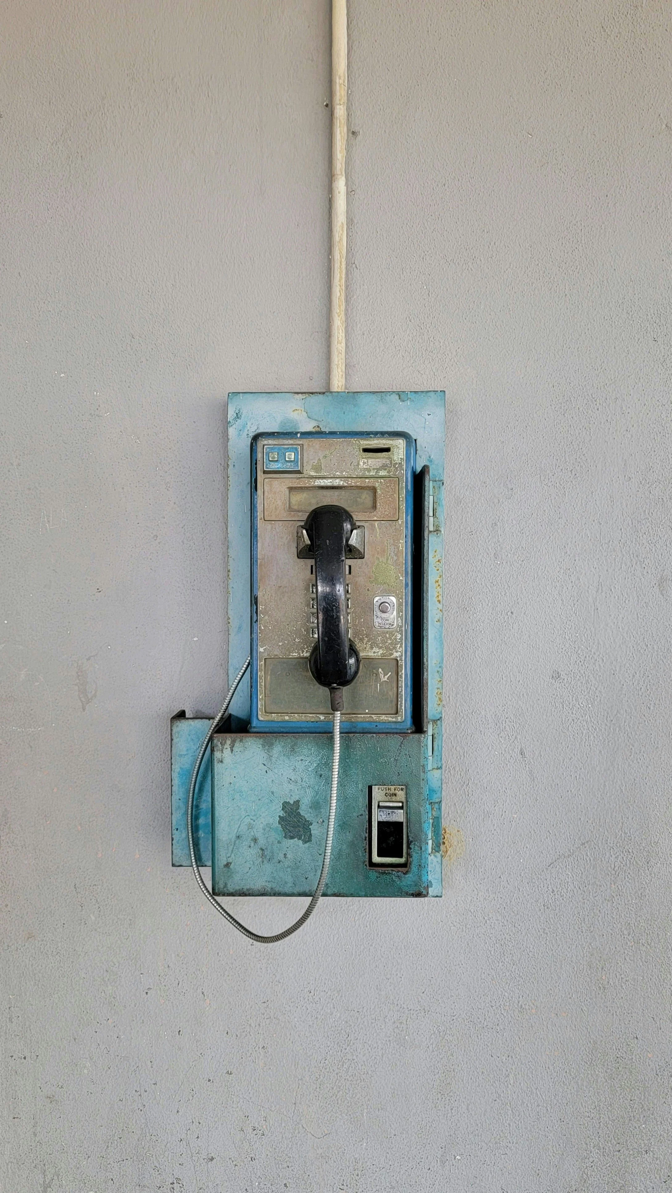 An old blue payphone mounted on a gray wall with a black handset hanging on the hook and a metal cord, featuring a coin slot and keypad area.