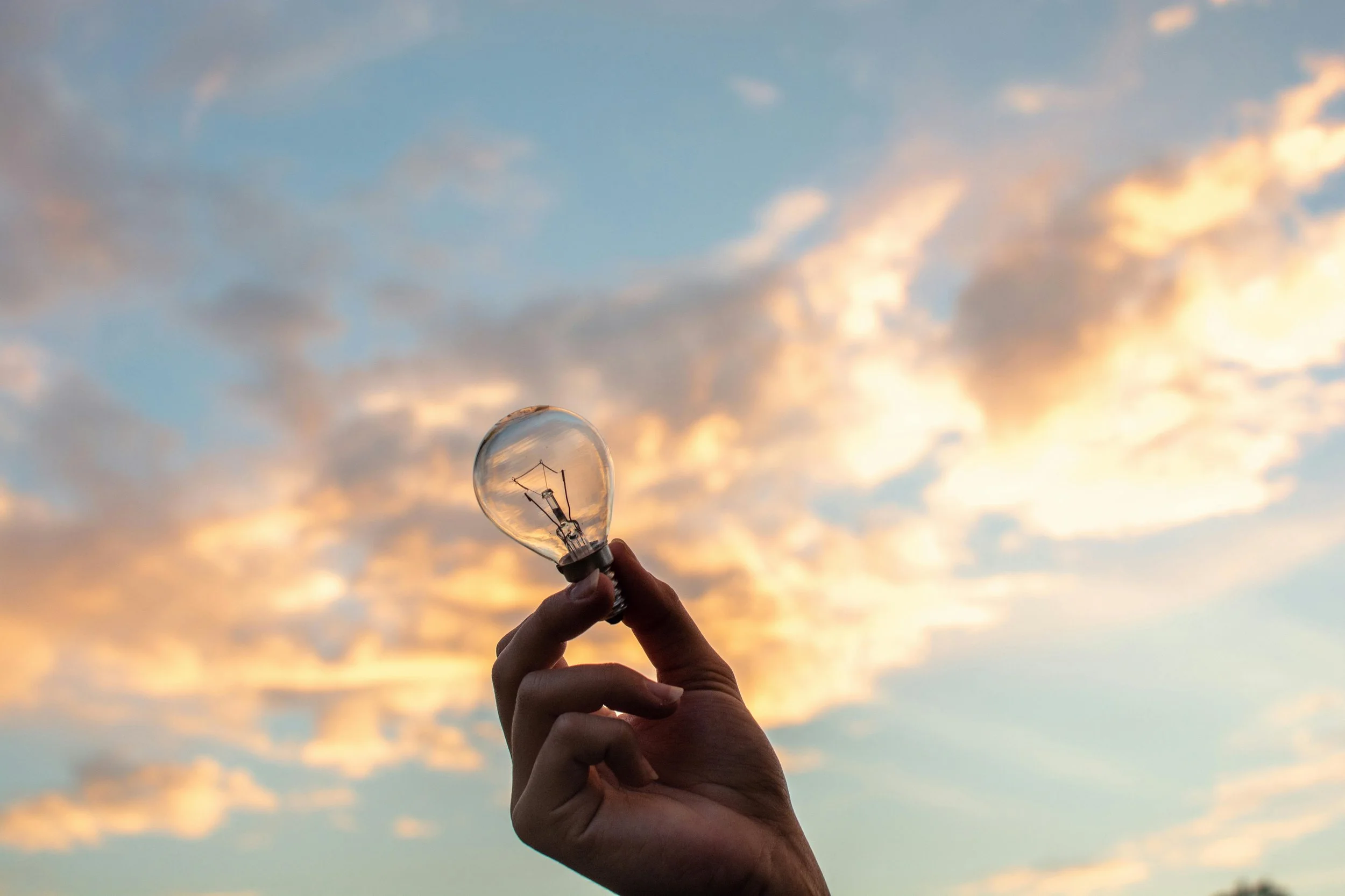 A hand holding a clear light bulb against a sky with orange and blue clouds at sunset.