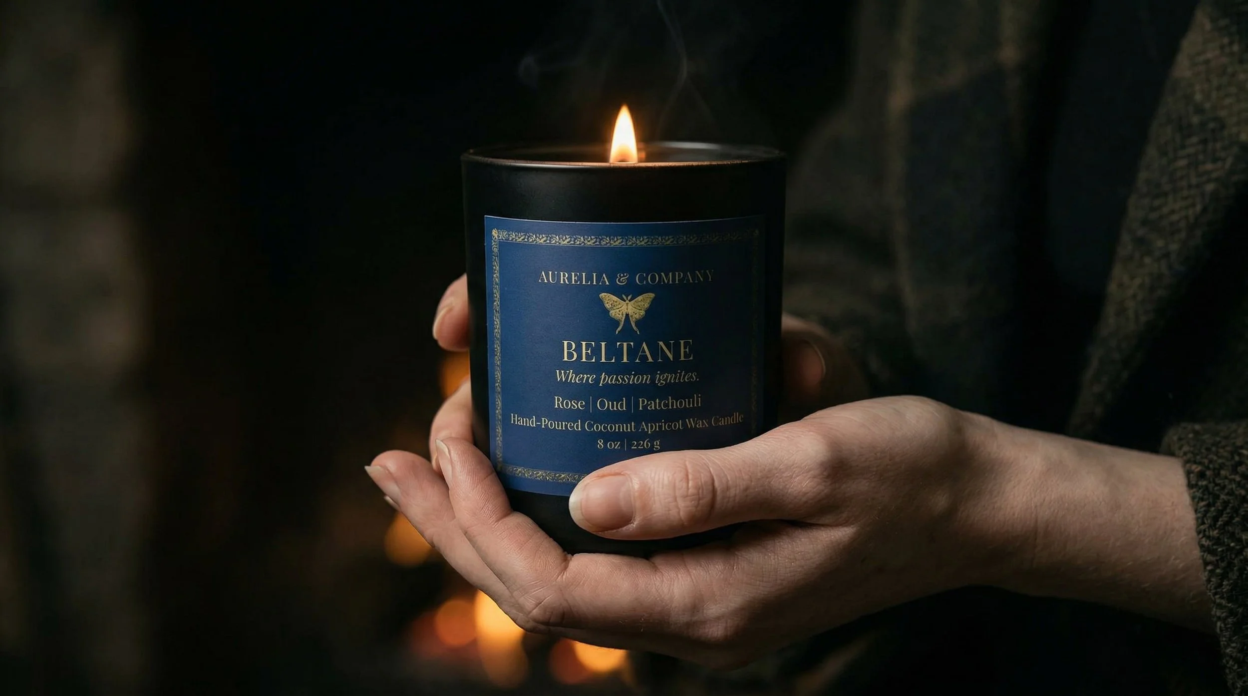 A person holding a lit candle with a blue label that reads "BELTANE" and features a butterfly logo. The candle is a hand-poured coconut apricot wax candle with scents of rose, oud, and patchouli.