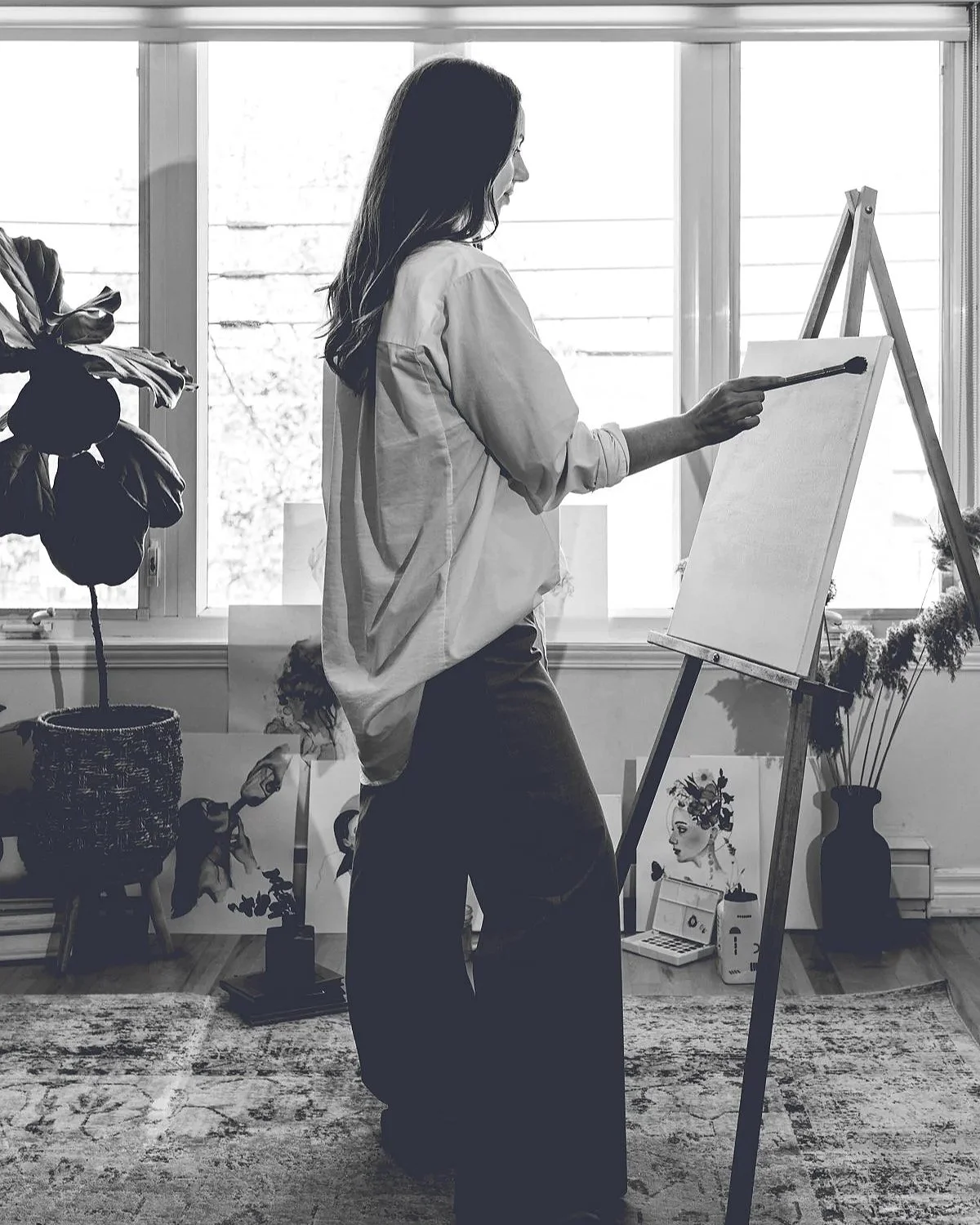 Une femme peignant sur une toile dans un studio lumineux avec des plantes et des œuvres d'art en arrière-plan.