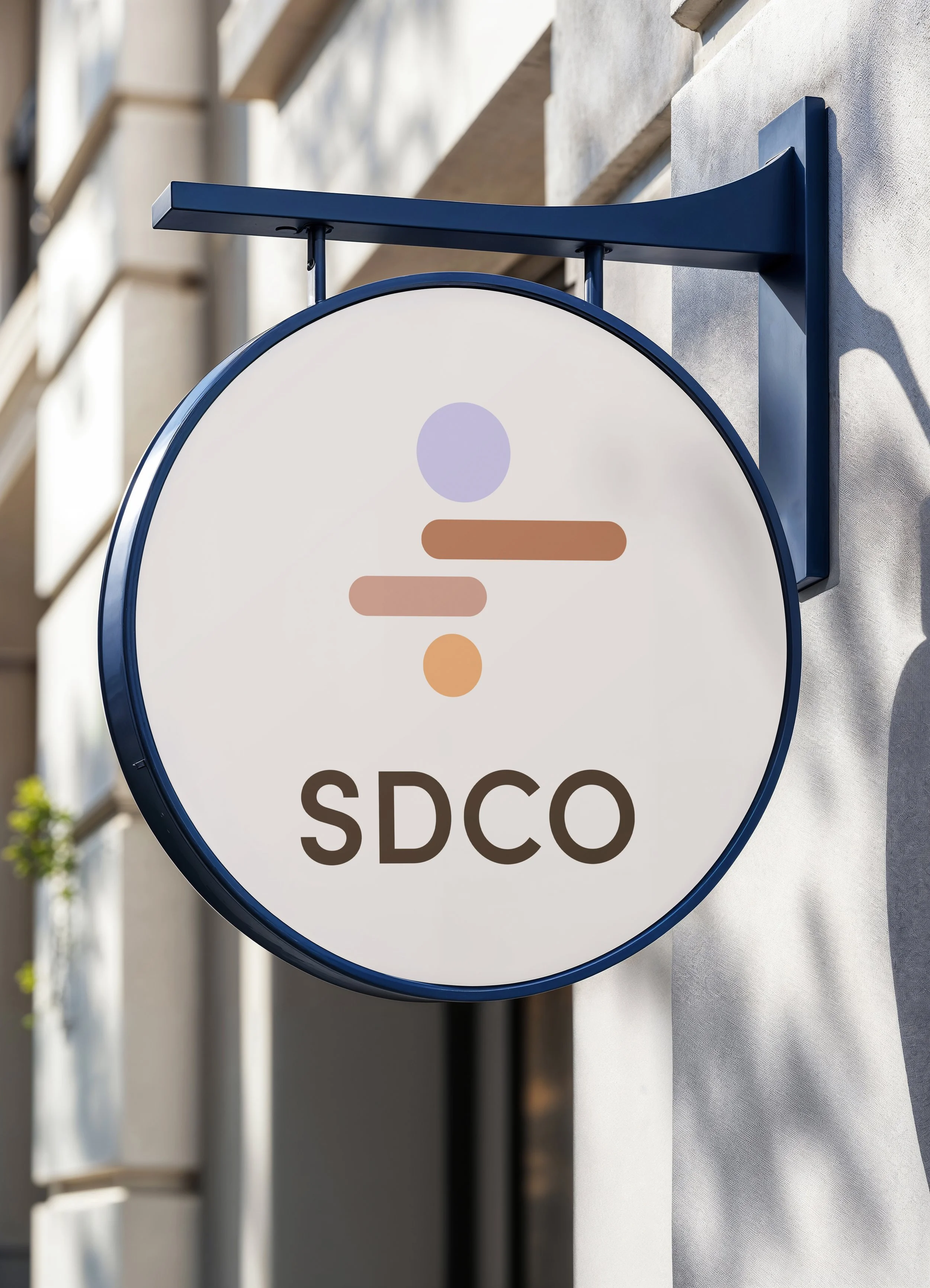 Signe rectangulaire blanc avec une icône de personne stylisée en haut et le texte « SDCO » en bas, fixé à un mur extérieur avec une structure métallique bleue.