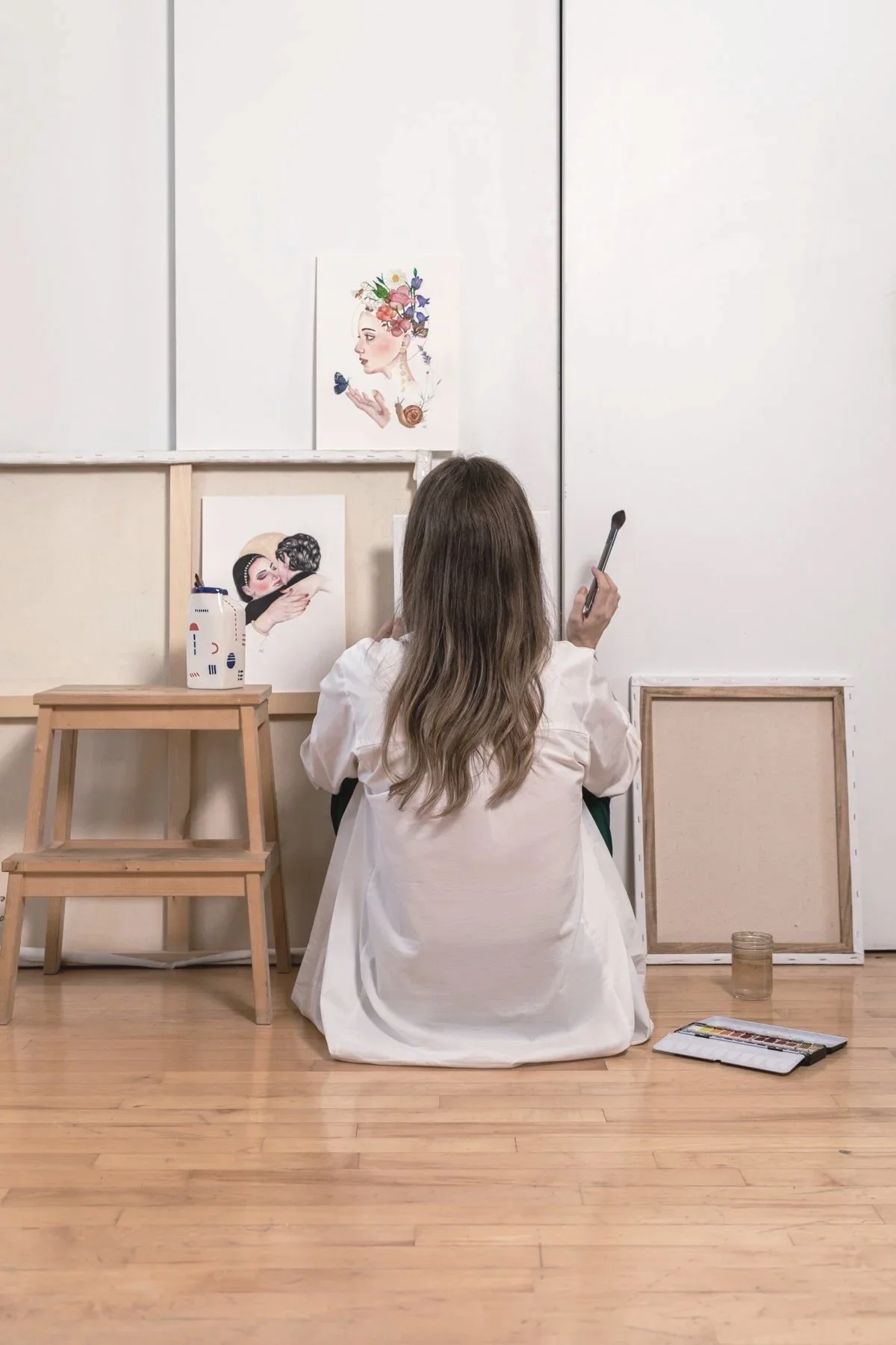Une femme vue de dos, assise par terre, avec des cheveux longs, mèche ondulée, portant une chemise blanche. Elle est dans une pièce avec un sol en bois, entourée de cadres vides et d'outils de peinture, regardant des œuvres d'art modernes sur un mur blanc