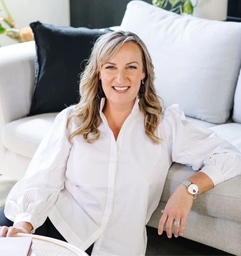 Kath Harris wearing a white blouse, sitting on a light-colored sofa with dark and light pillows behind her, in a cozy living room setting.