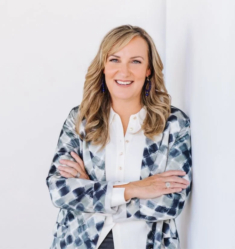 Kath Harris wearing a white blouse, a gray and blue patterned blazer, and blue earrings, standing with arms crossed against a white wall.