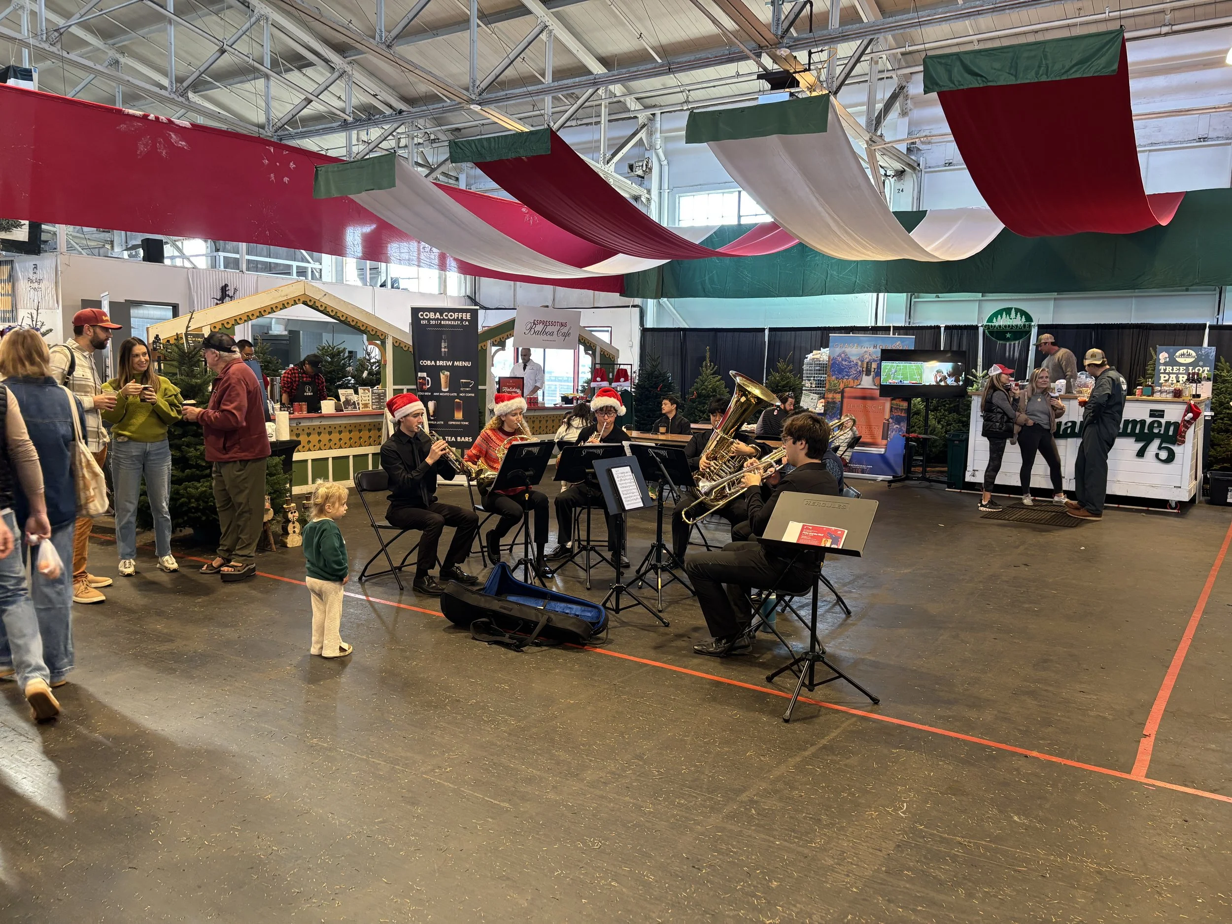 A holiday indoor market with a live band wearing Santa hats, Christmas trees, and festive decorations, surrounded by shoppers.
