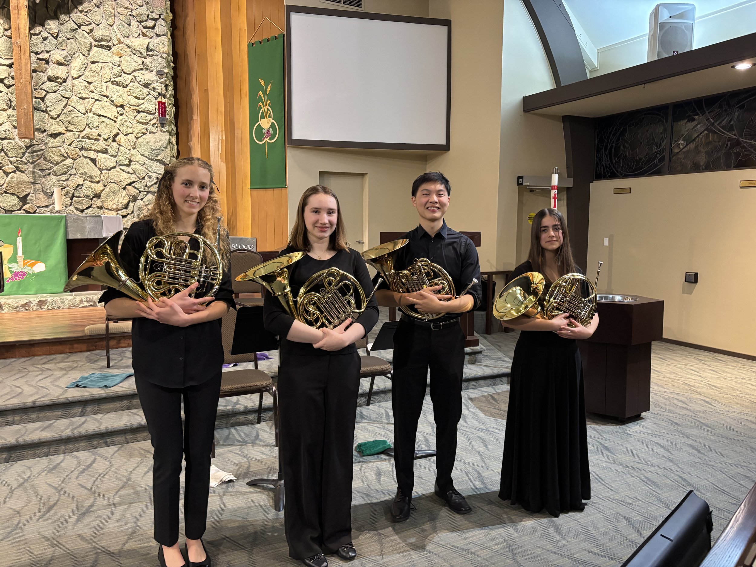 Four young musicians in formal black attire holding French horns standing in a church or performance venue.