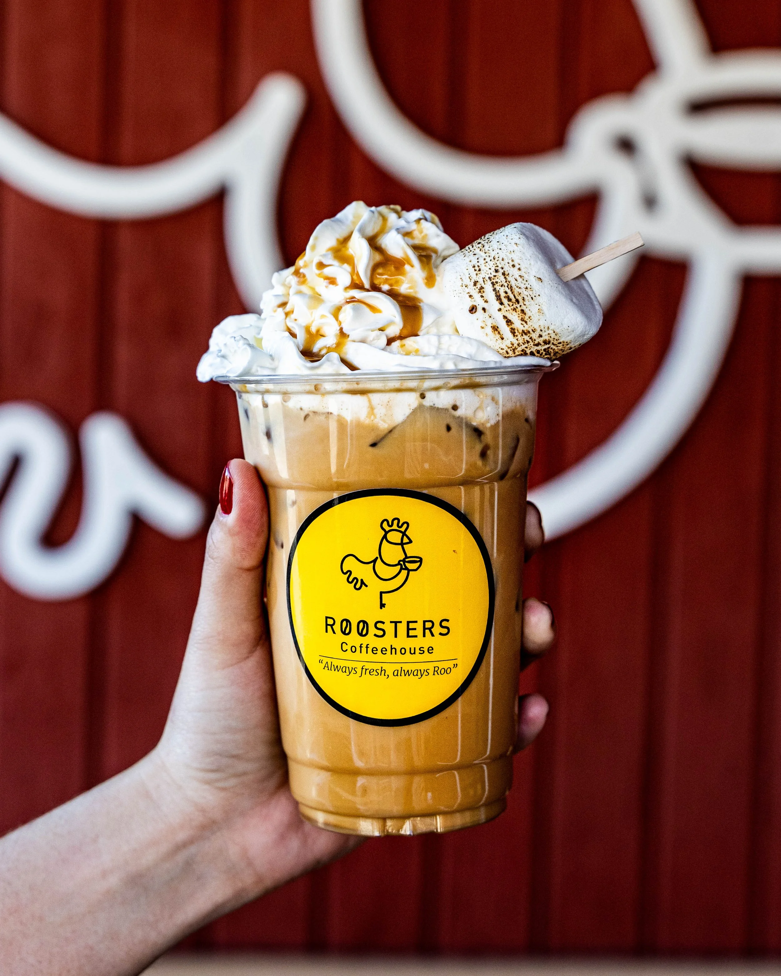 A hand holding a large iced coffee with whipped cream, caramel drizzle, and toasted marshmallow on top, in front of a red wooden background with a white rooster logo.