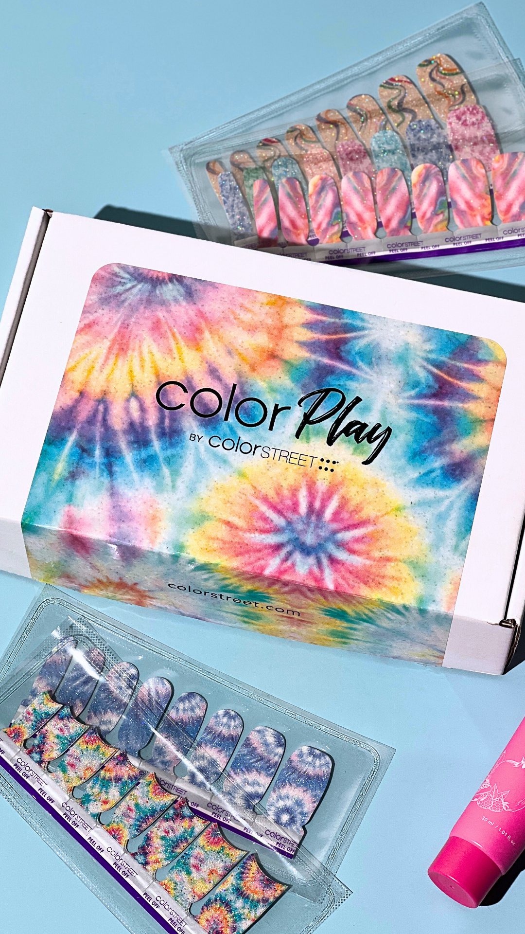 A box of tie-dye nail wraps with colorful patterns, a set of nail wraps in a plastic case, and a pink bottle of lotion or moisturizer on a blue surface.