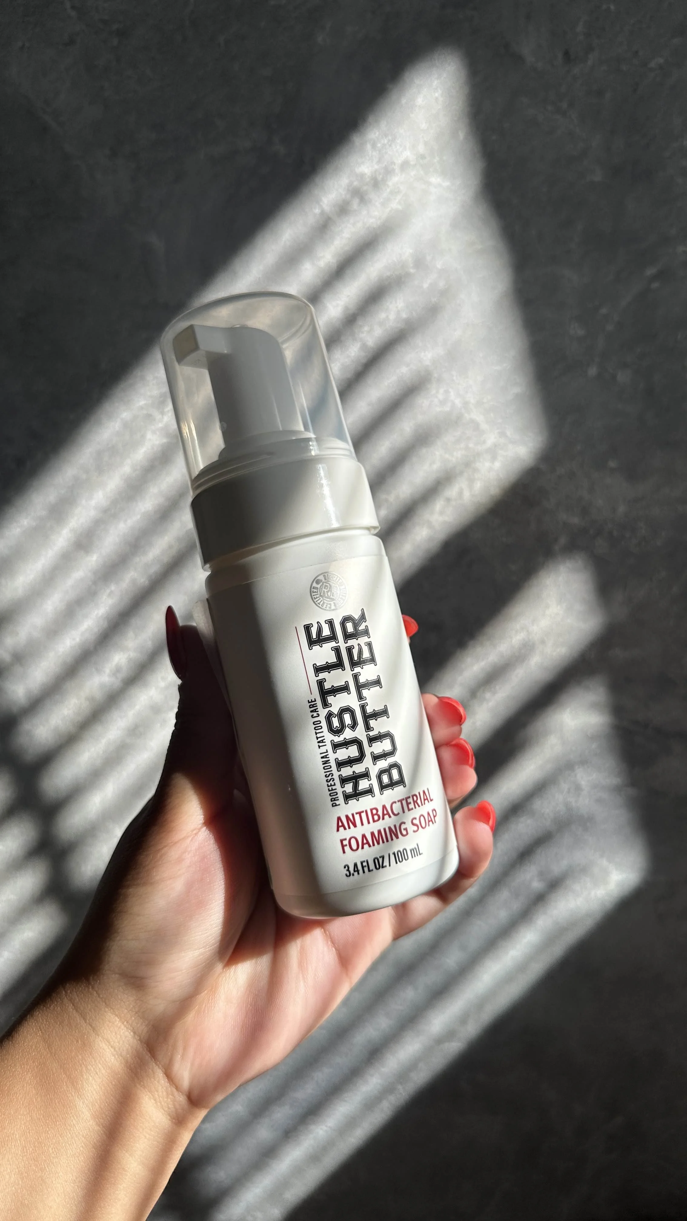 Hand holding a bottle of Hustle Butter antibacterial foaming soap with shadows of blinds cast on the surface behind.