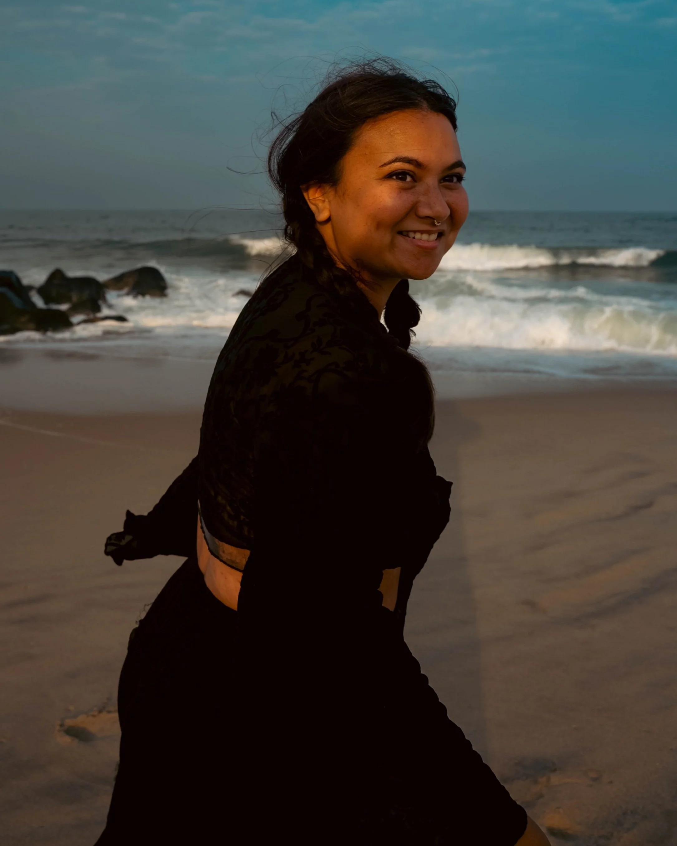 A woman with dark hair tied in braids, smiling, sitting on a sandy beach with ocean waves and rocks in the background.