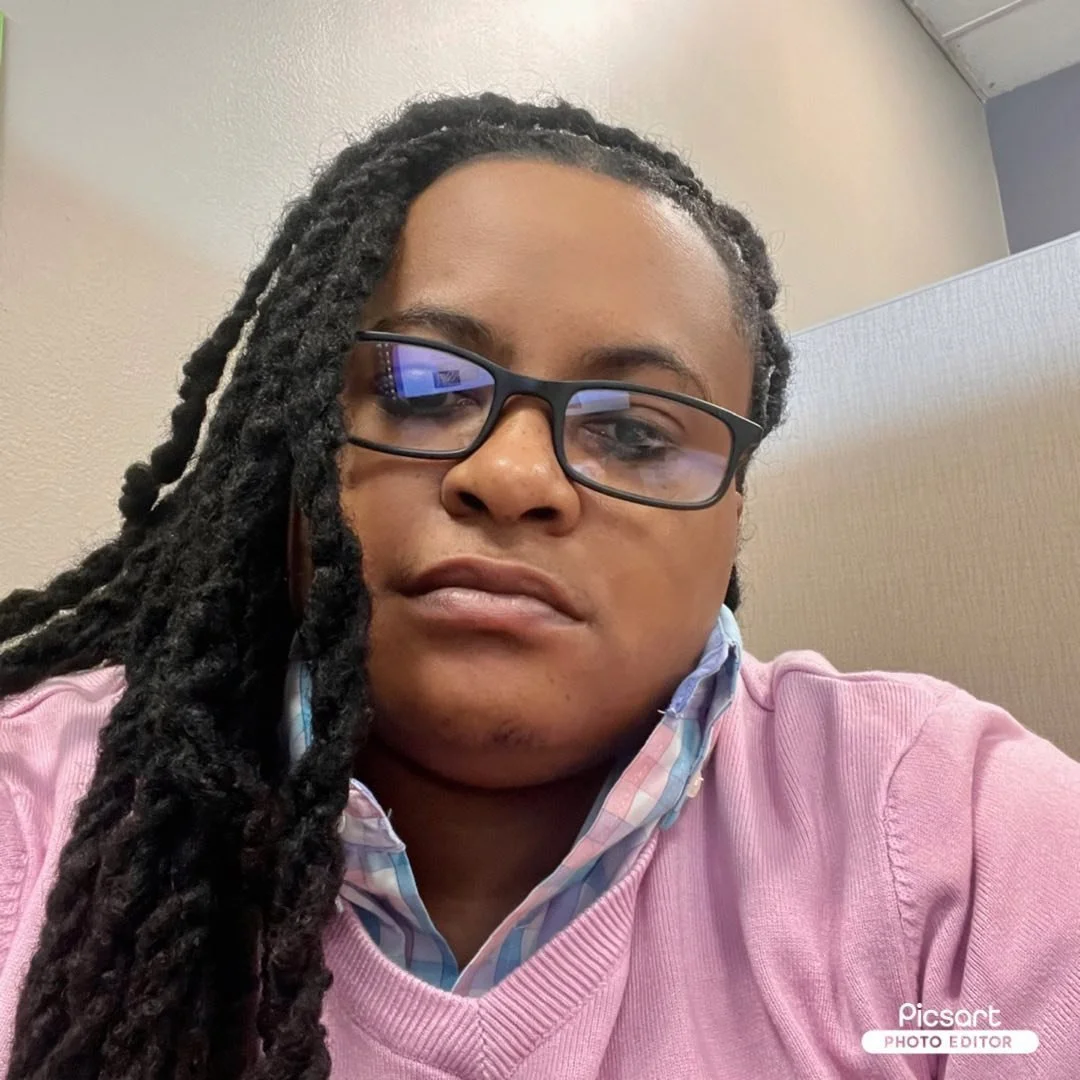 A woman with glasses and long dreadlocks, wearing a pink sweater over a plaid shirt, sitting against a beige wall.