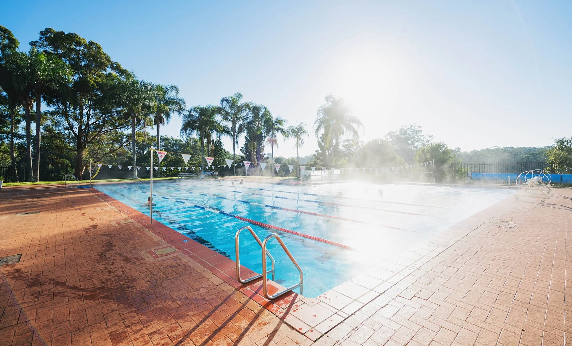 Outdoor swimming pool with lane markers and steam rising from the water, surrounded by trees and clear sky.