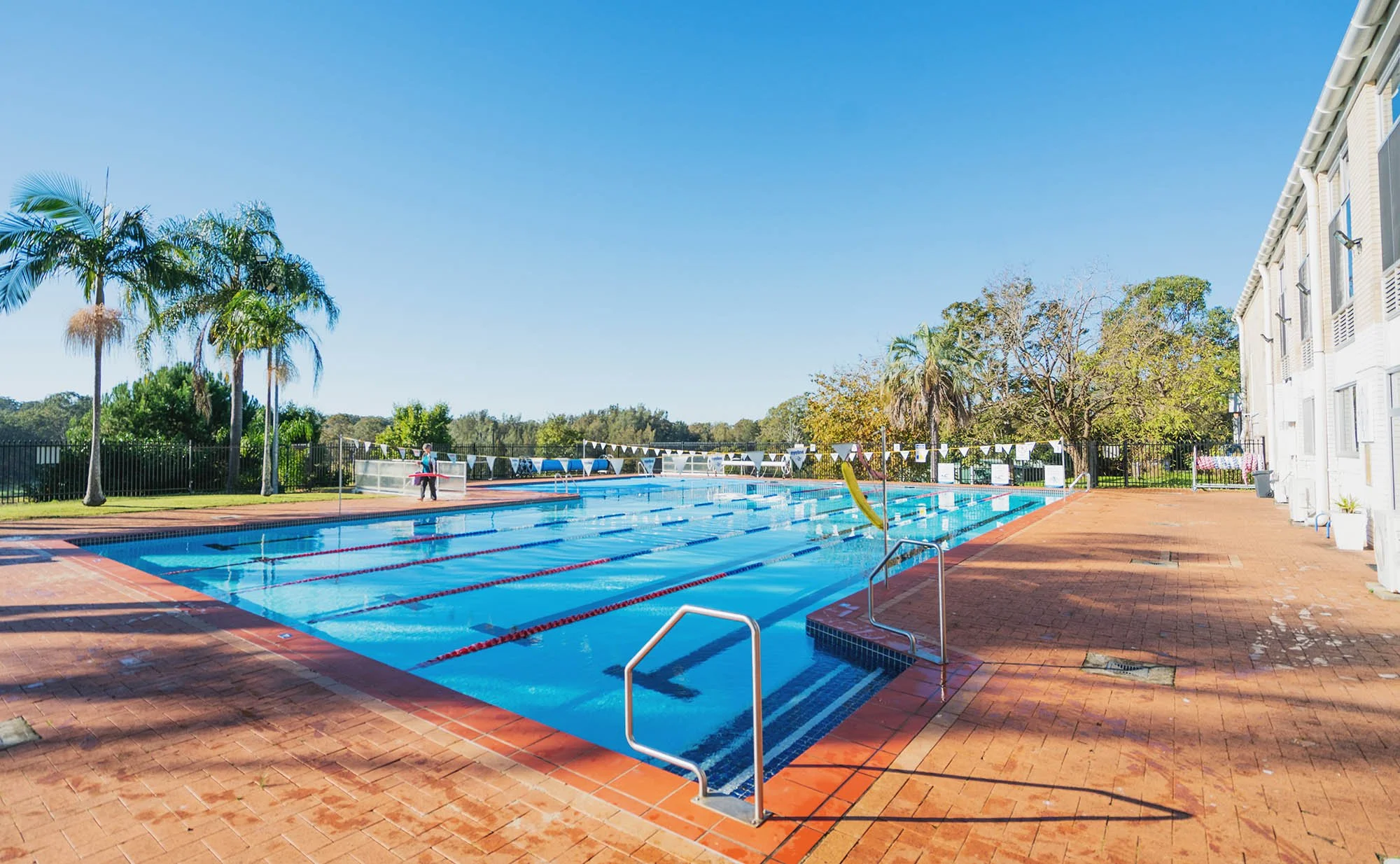 Empty outdoor swimming pool with lanes, surrounded by orange brick pavement, palm trees, and a white building during daytime with clear blue sky.